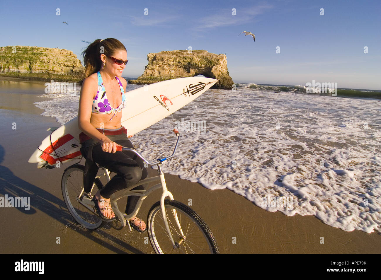 Woman Riding Cruiser A woman with a surfboard riding a cruiser bike on the beach at Natural ...