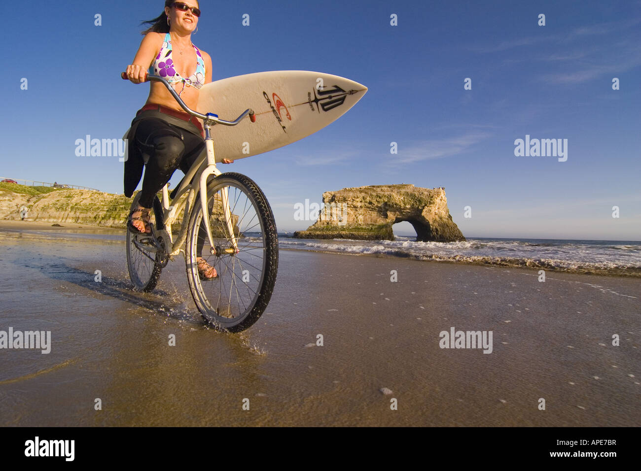 Woman Riding Cruiser A woman with a surfboard riding a cruiser bike on the beach at Natural ...