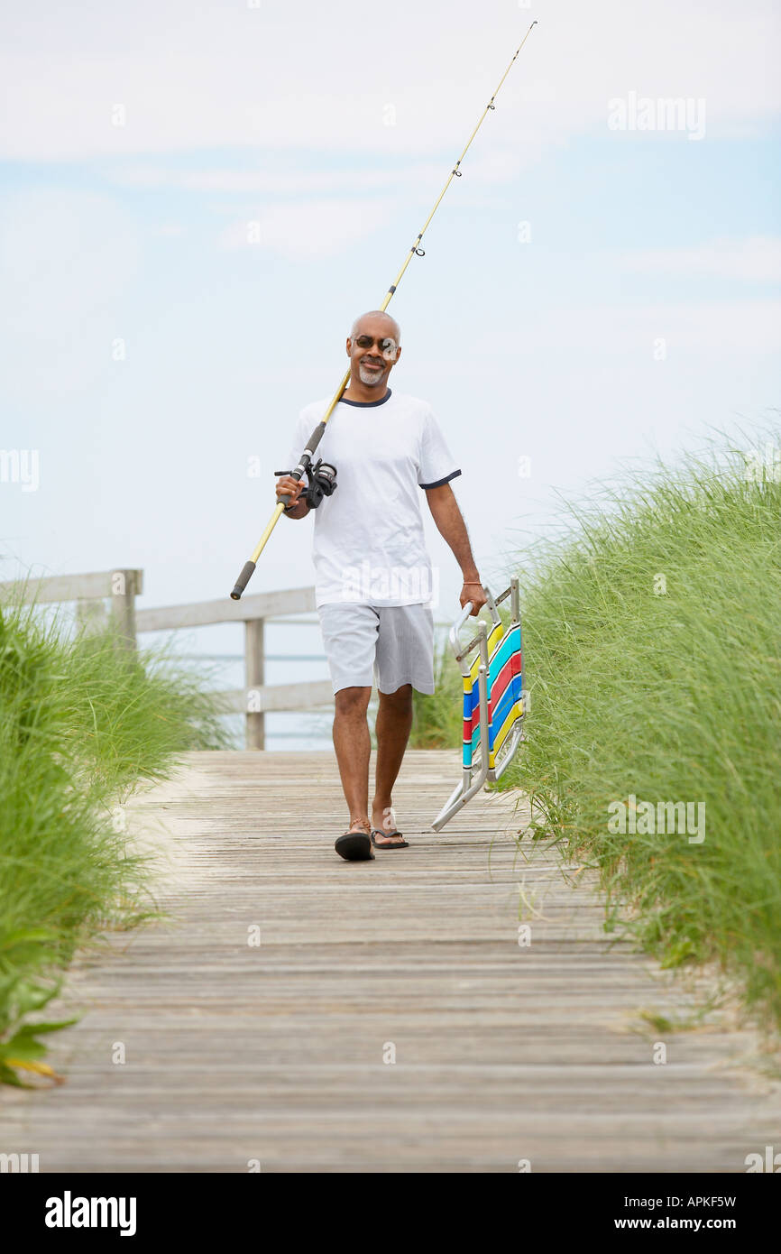 Man with fishing rod and folding chair walking on boardwalk Stock