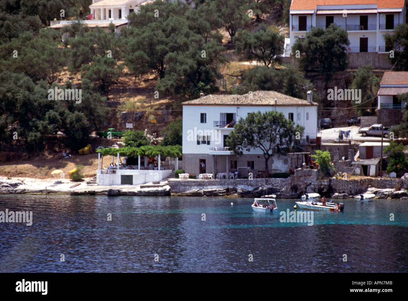 A few pleasure boats moored in the clear waters in front of the pebble beach of Kalami on the island of Corfu a tree in front of Lawrence Durrell s White House where he wrote Prospero s Cell and The White House beyond Stock Photo