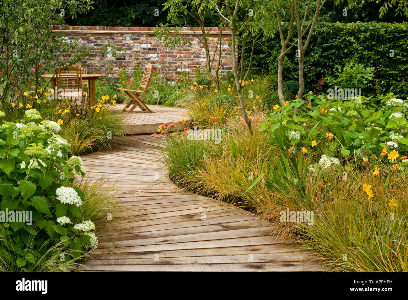 Boardwalk in garden to decking sitting area with table and chairs. Plants include Hydrangea Hemerocallis evergreen grass Stipa a Stock Photo