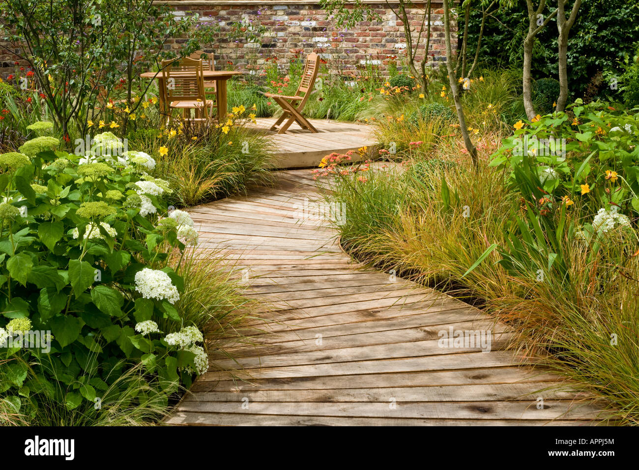 Boardwalk in garden to decking sitting area with table and chairs. Plants include Hydrangea Hemerocallis evergreen grass Stipa Stock Photo