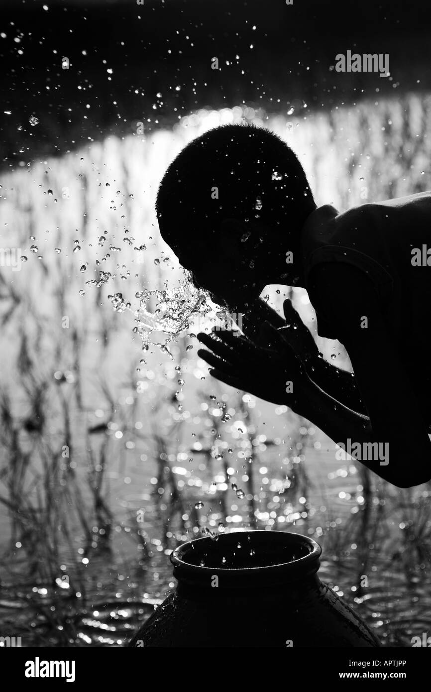 Silhouette of a rural Indian village boy face washing from a clay pot next to of a rice paddy field. Andhra Pradesh, India. Black and white Stock Photo