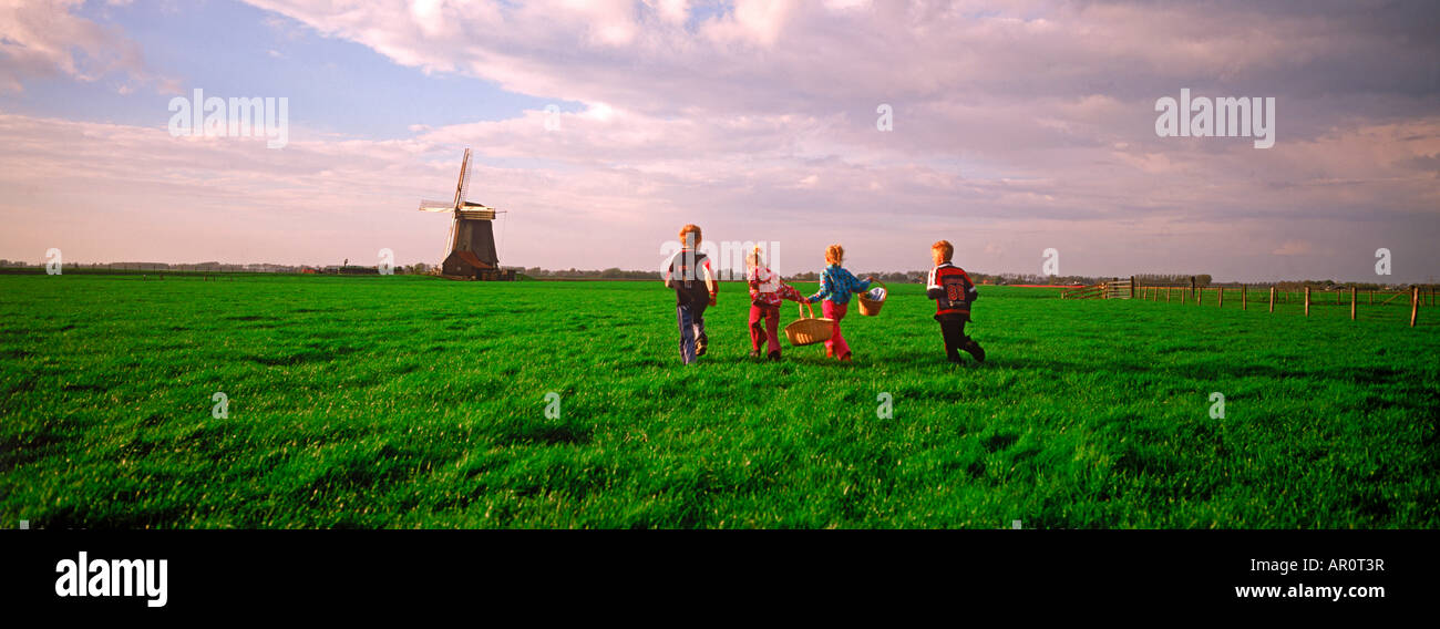 Four children with baskets crossing farmlands in Holland near windmill at sunset Stock Photo