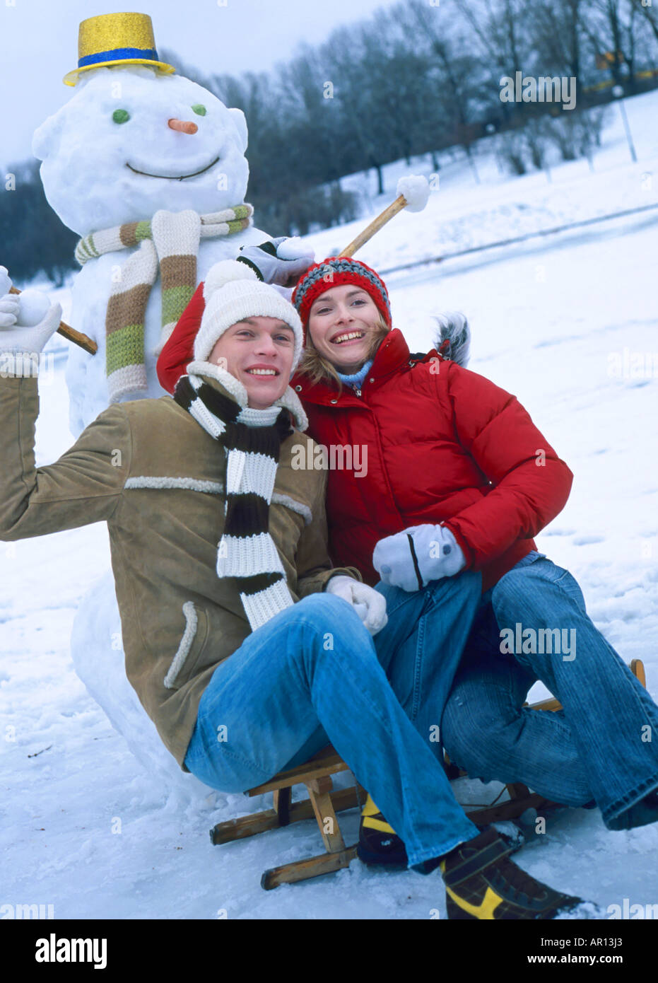outdoor day winter snow tree trees park woman man 25 30 young pair couple smile smiling hug embrace crouch cap pompom caps sc Stock Photo