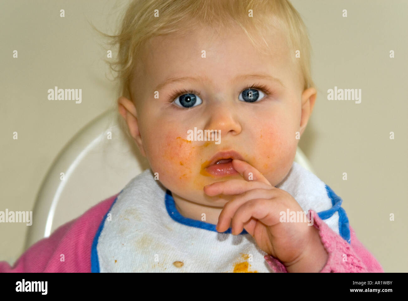 Horizontal close up of a pretty Caucasian baby girl sitting in a high chair at dinner time with food all over her face and hands Stock Photo