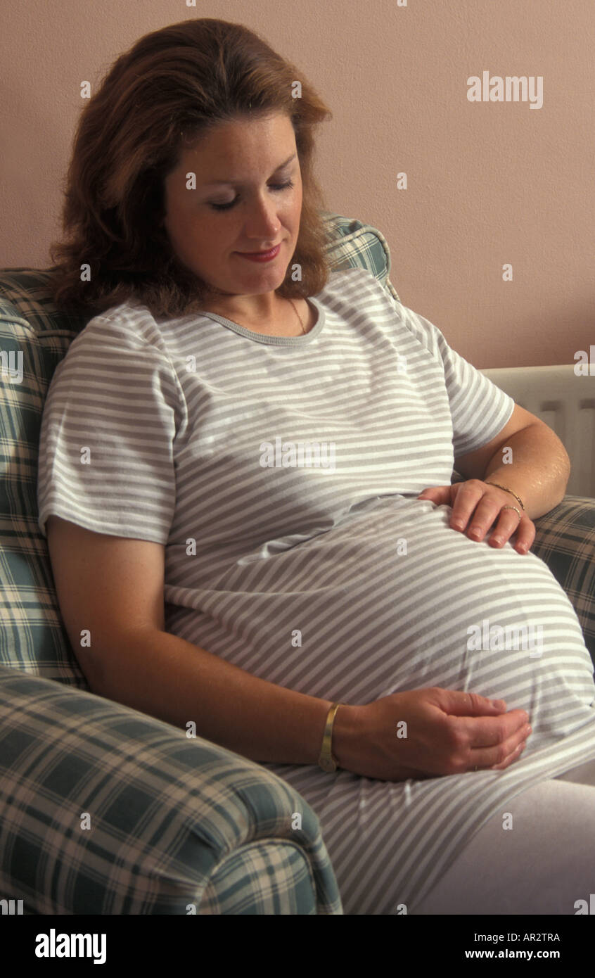 heavily pregnant caucasian woman in her thirties sitting in armchair looking down at tummy Stock Photo