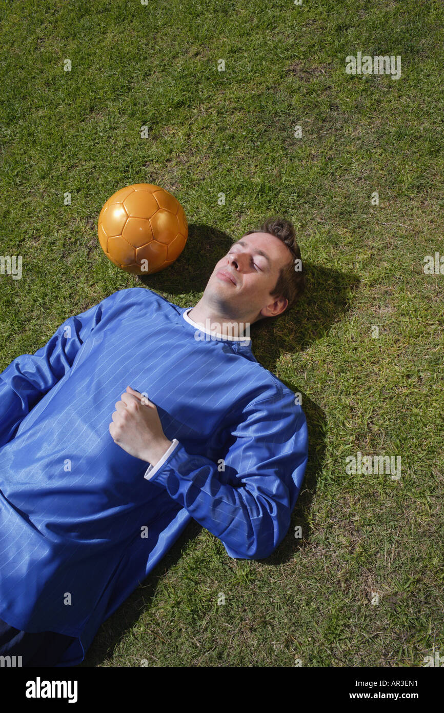 Young man wearing football shirt lying on grass next to a golden coloured football Stock Photo