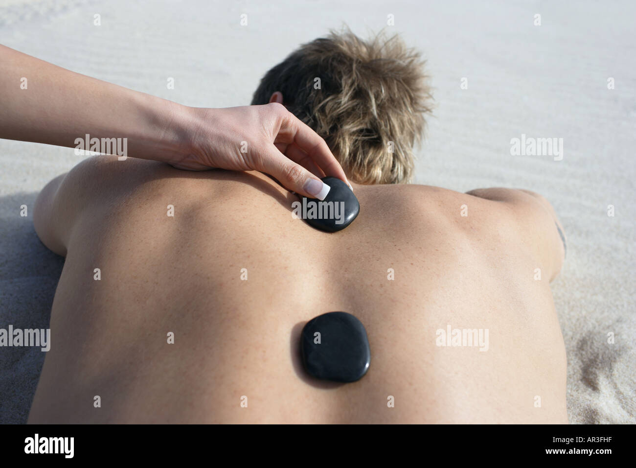 Young woman placing black hot stones on man's back lying on beach Stock Photo