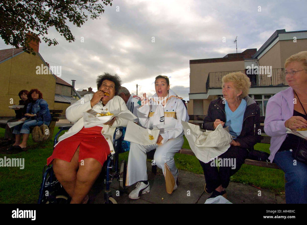 Female Elvis impersonator eating fish and chips with friends on a bench during the annual Elvis Festival in Porthcawl, UK. Stock Photo