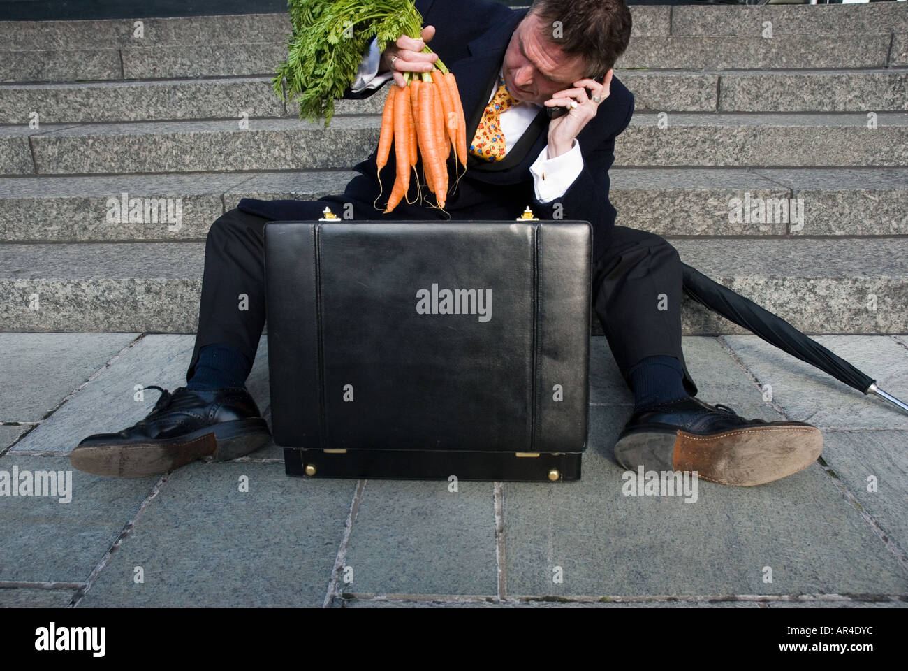 Bussiness man on mobilephone withbriefcase,sittng down on steps,holding a bunch of carrots Stock Photo