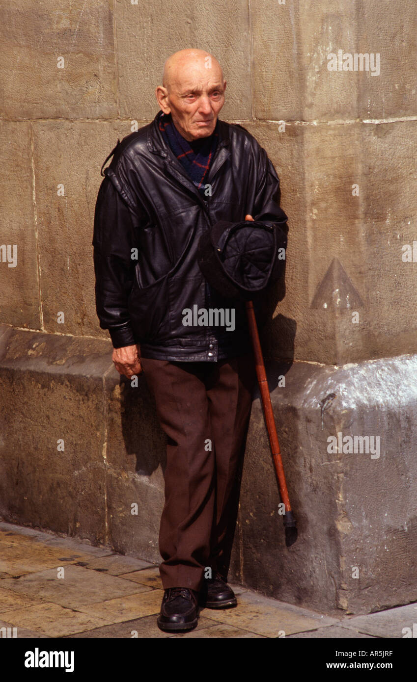An elderly man leaning against a stone wall while holding a walking cane and a cap in Krakow Poland Stock Photo