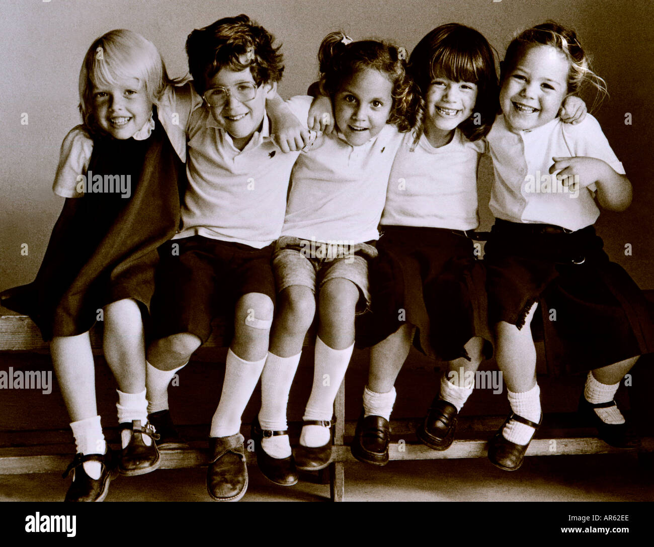 B&W Retro school 1980’s group of happy junior school children in uniform sitting arms around each other on bench in school classroom retro B&W Stock Photo