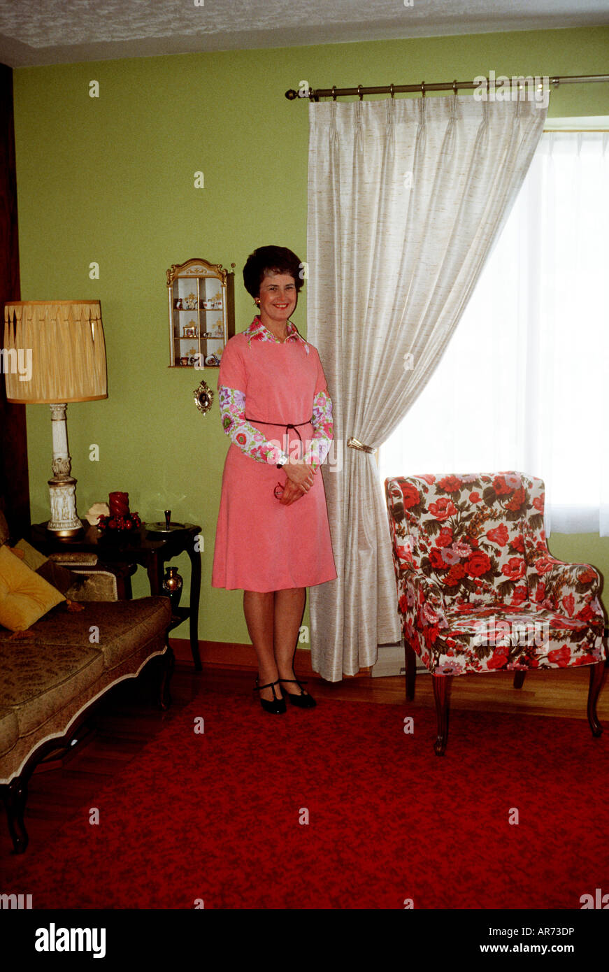 70s housewife in pink dress stands proudly in the corner of red and yellow living room. Patterned chair and ornate couch. Stock Photo