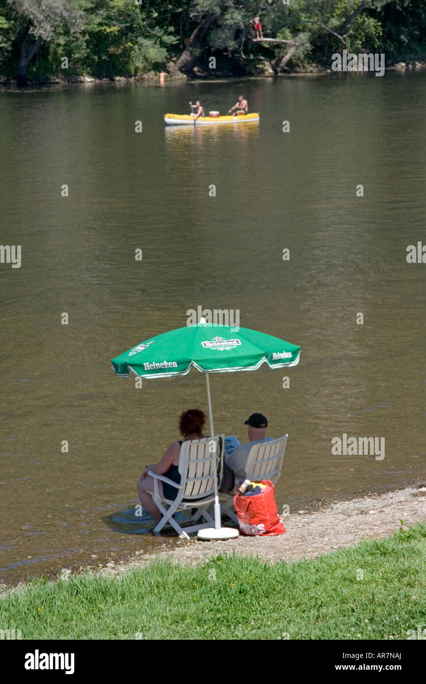 Couple relax under the shade of a parasol at the edge of a river whilst a couple paddle past in a canoe in the background. Stock Photo