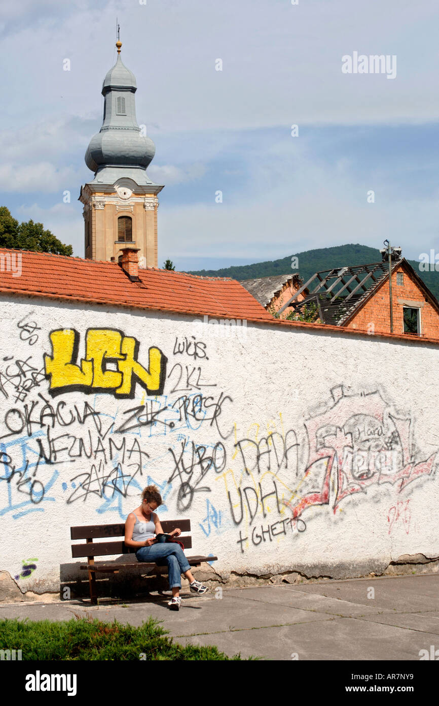 A young lady reading on a bench in front of a graffiti covered wall in the town of Roznava in eastern Slovakia. Stock Photo