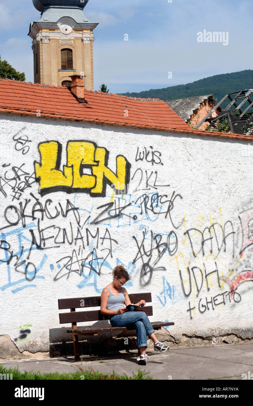 A young lady reading on a bench in front of a graffiti covered wall in the town of Roznava in eastern Slovakia. Stock Photo