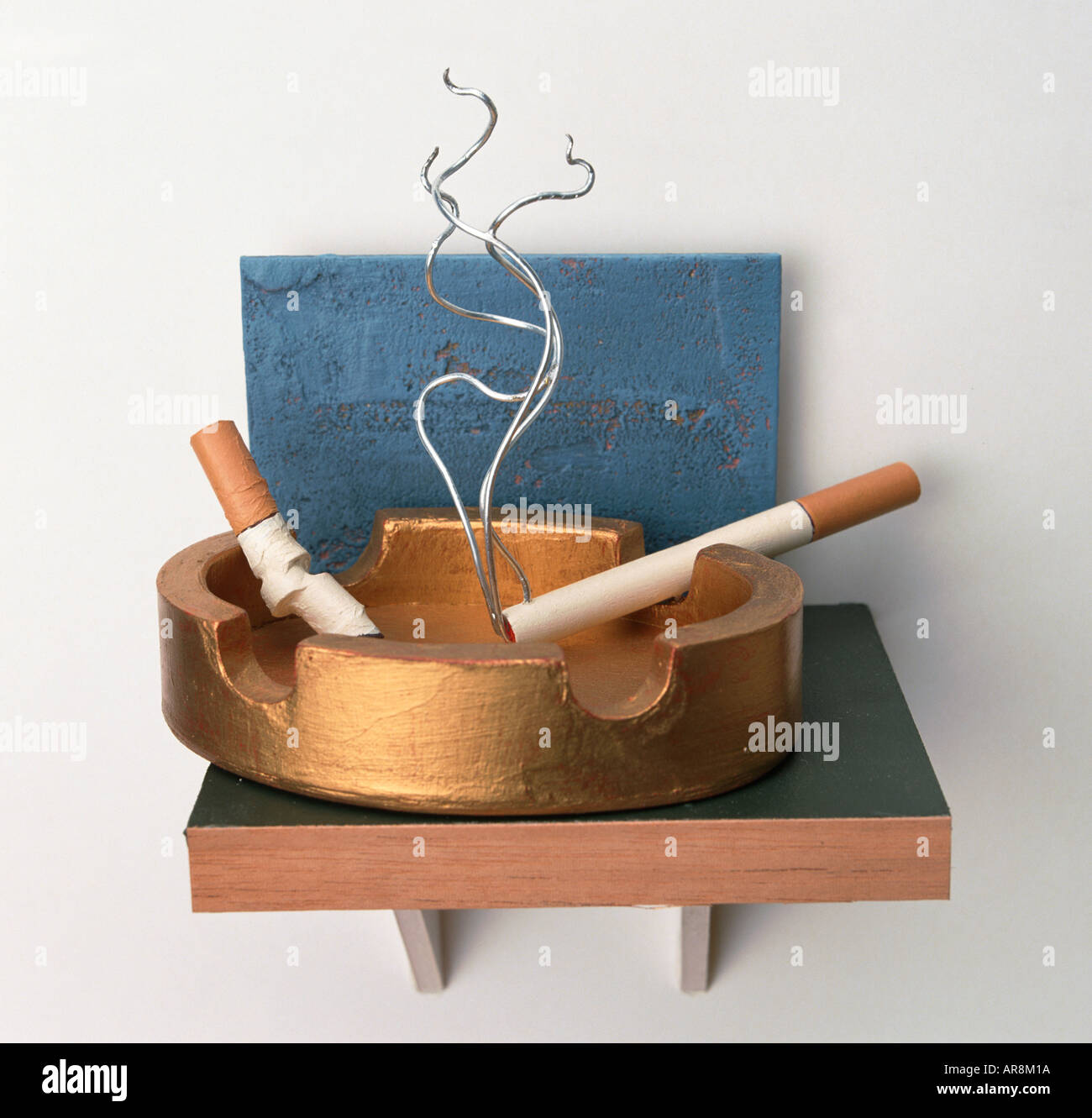 two cigarettes in an ashtray Two model cigarettes burning in golden ashtray, metal wires forming smoke  spiralling from one cigarette, blue background Stock Photo