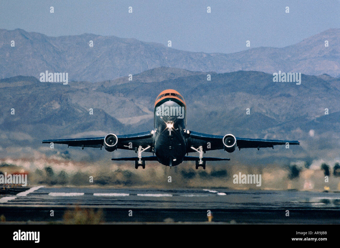 widebody jumbo jet airliner taking off just airborne above runway head on view with hills mountains in background Stock Photo