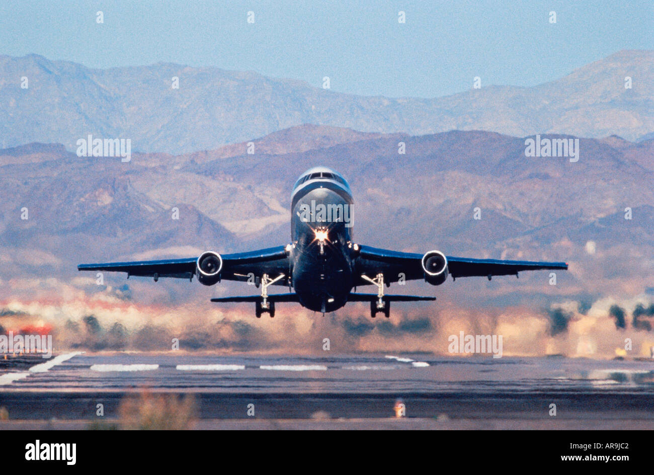 widebody jumbo jet airliner taking off just airborne above runway head on view with hills mountains in background Stock Photo