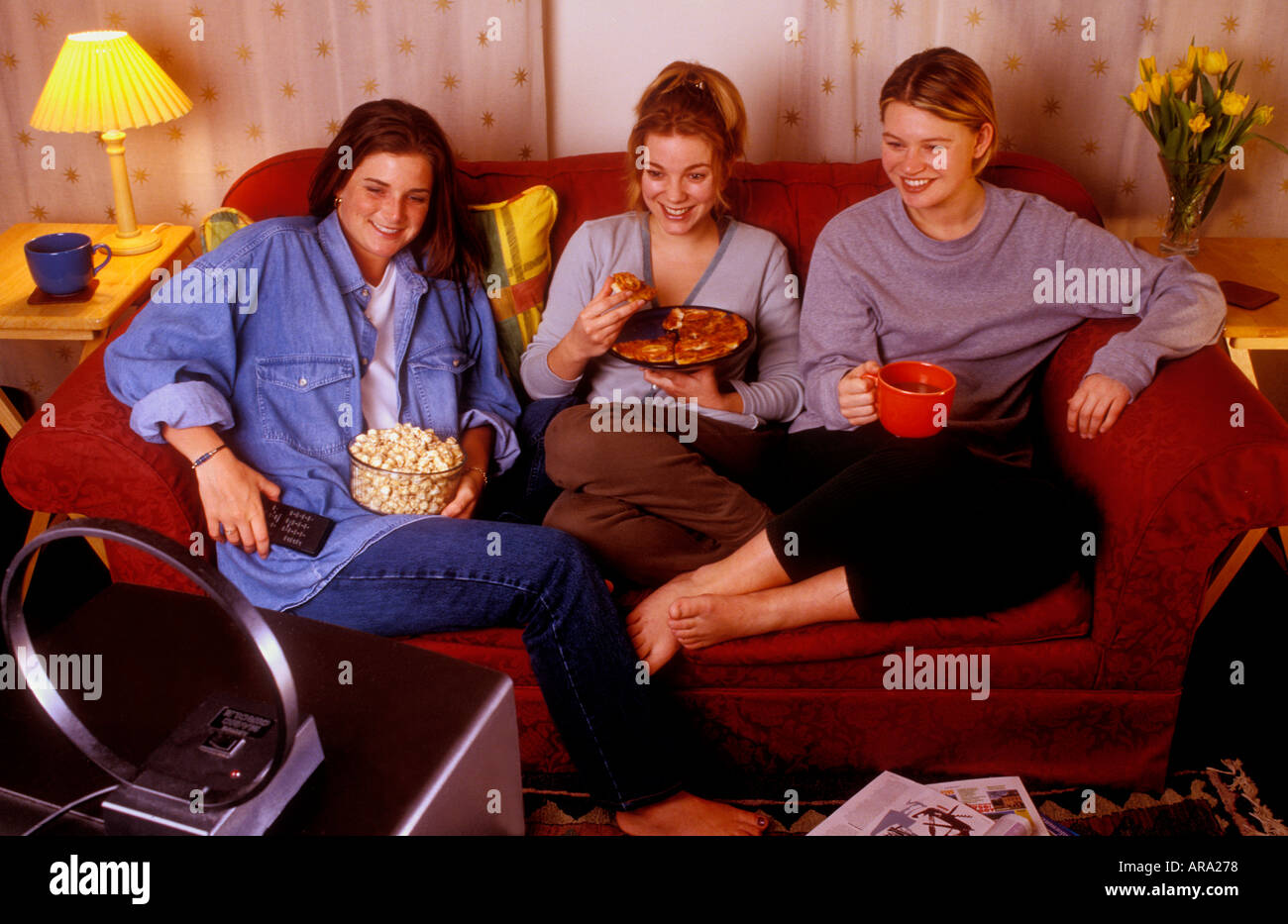 flatmates girls pizza popcorn snack sharing a sofa watching small portable TV in their shared rental home living room Stock Photo