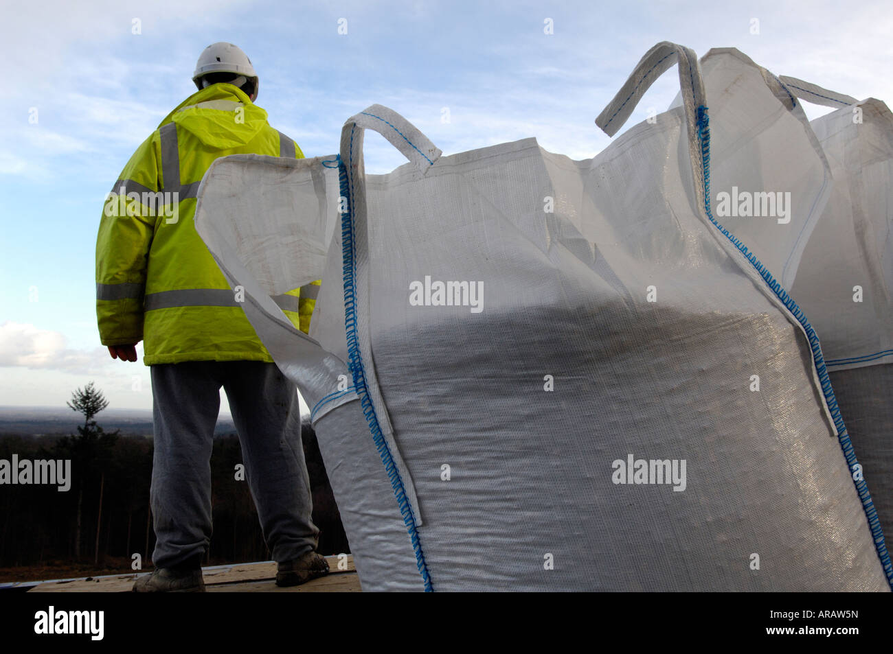 a builder workman stood on atrailer on a building site unloading large whit sacks bags of ballast and raw materials wearing ppe Stock Photo