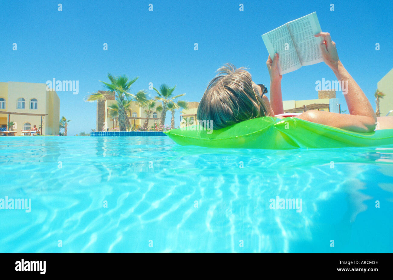 woman 40 floating on inflatable airbed on clear blue water swimming pool reading a book palm trees background Stock Photo