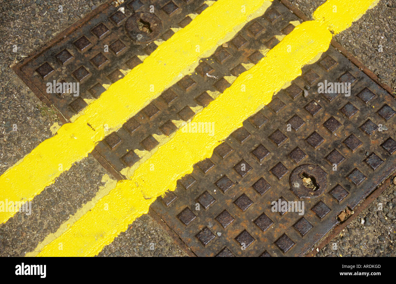 Diagonal detail from above of freshly painted double yellow lines crossing a square metal manhole cover set in tarmac road Stock Photo