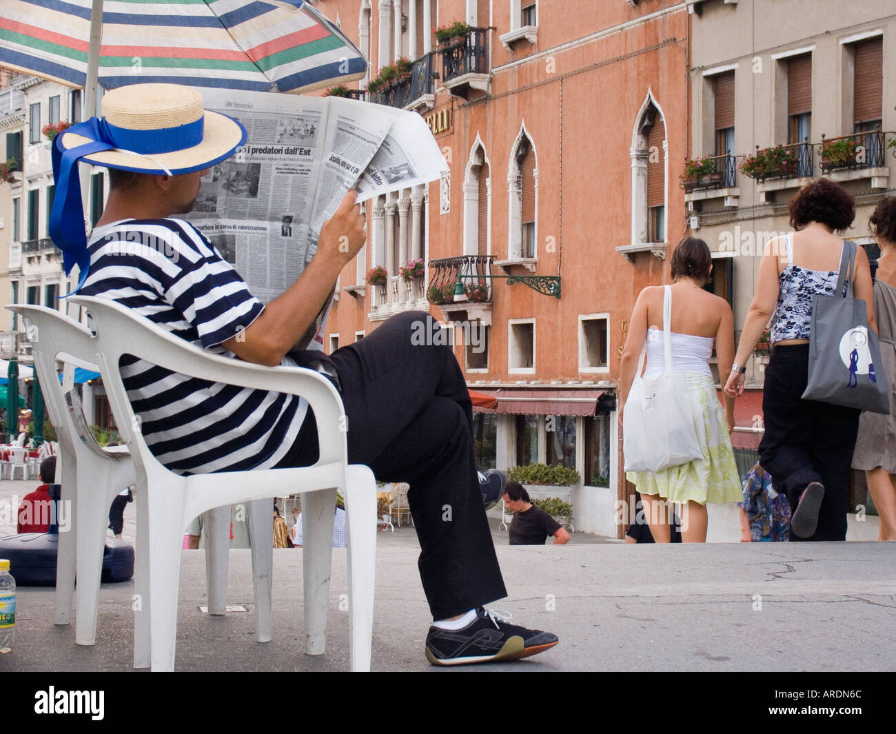 Gondolier wearing traditional straw hat and striped shirt sits reading newspaper while waiting for next customer Venice Italy Stock Photo