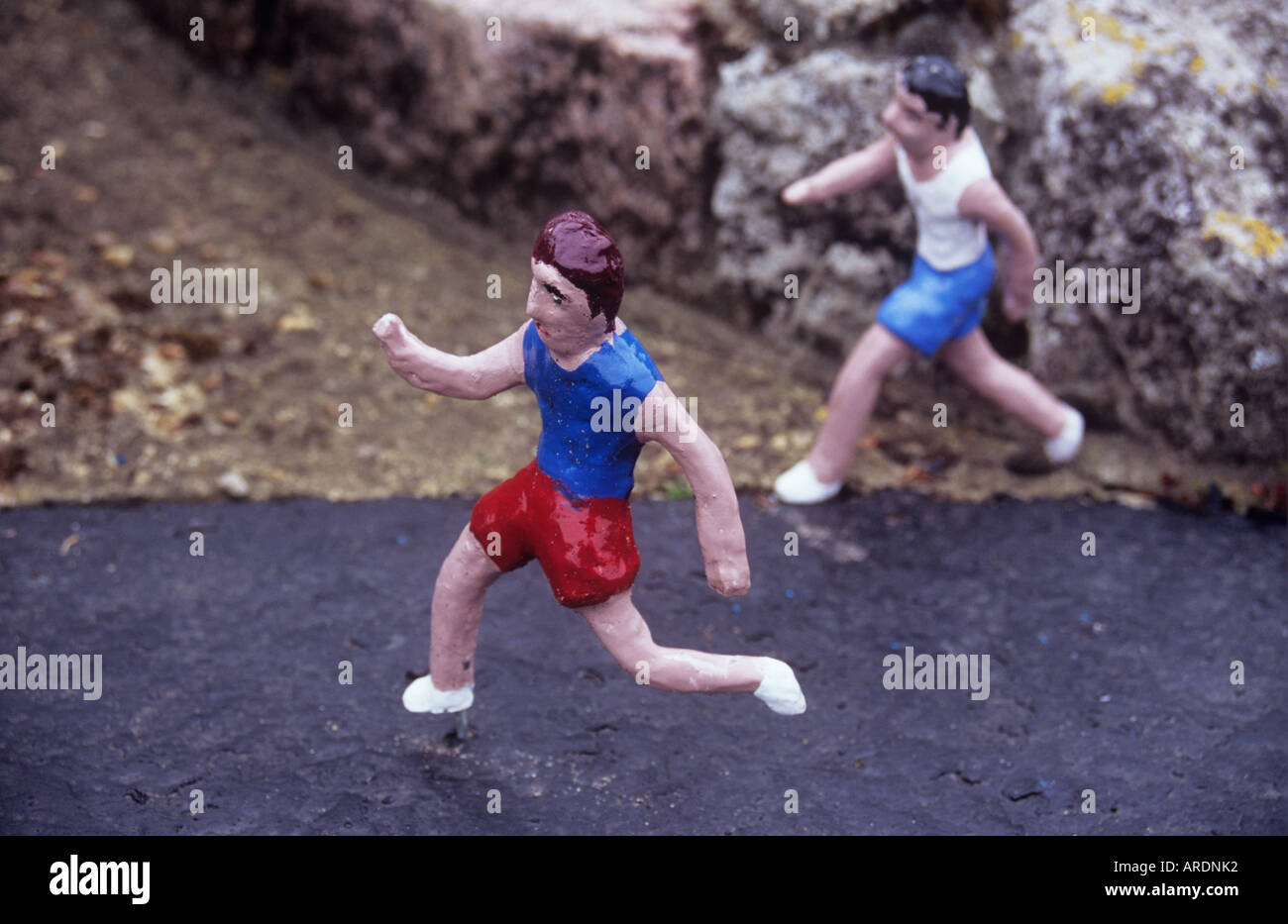 Two naively fashioned clay models painted in running gear striding out on miniature road past some rocks Stock Photo