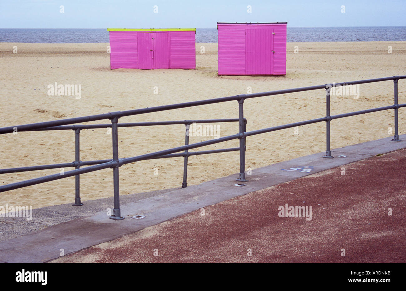 View from sandblown red asphalt promenade across railings to deserted dry white sandy beach with two pink sheds perched on it Stock Photo