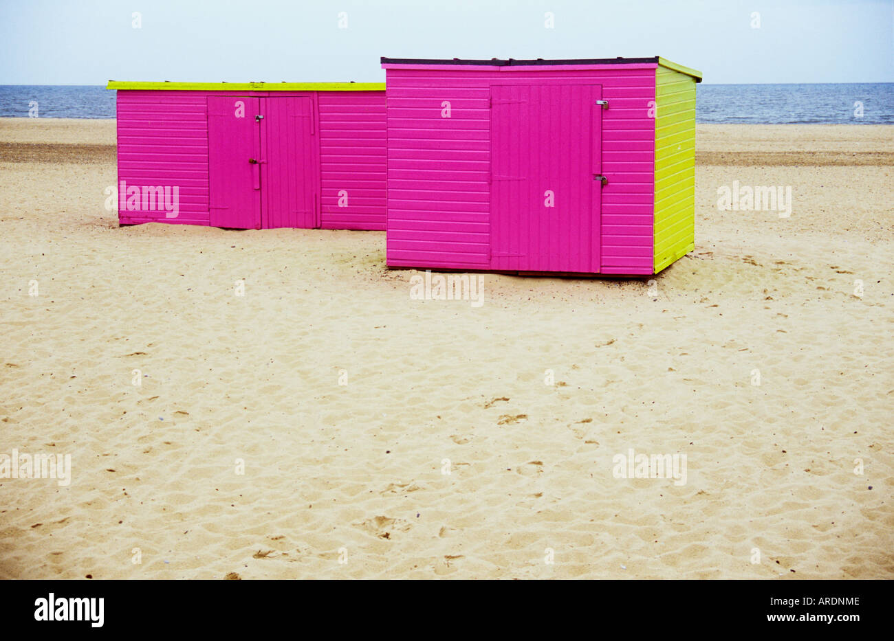 View of deserted dry white sandy beach with footprints and two pink and lime green sheds perched on it Stock Photo