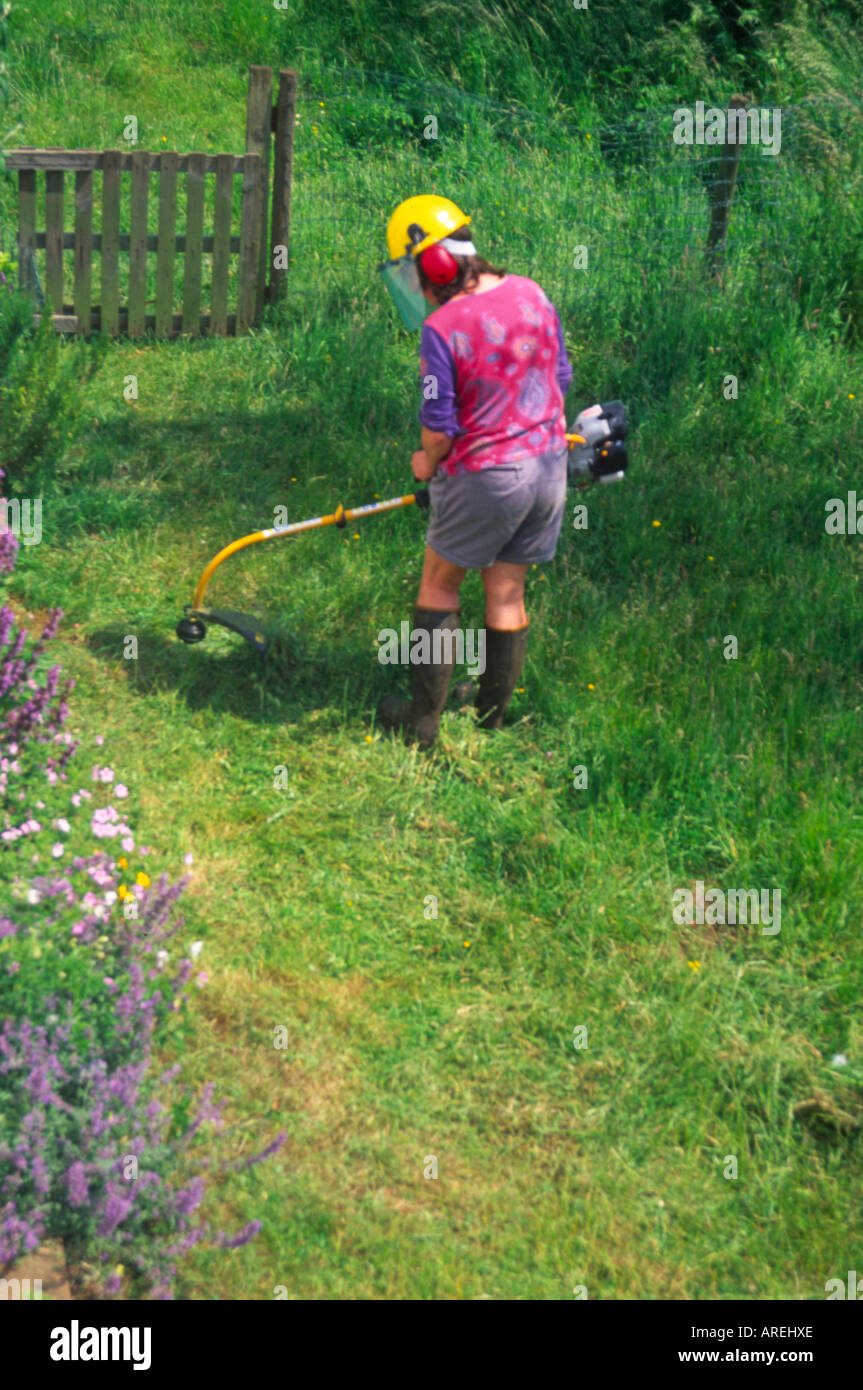 Woman using a strimmer to cut long grass lawn in her cottage garden