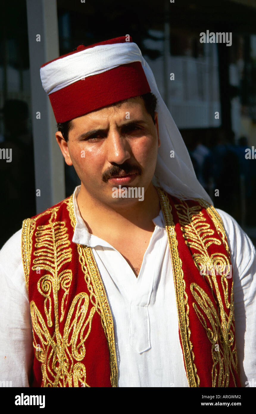 A serious looking gentleman models the traditional Turkish costume of a red  hat encircled with a white scarf which trails over his shoulder and a red  embroidered waistcoat worn over a white