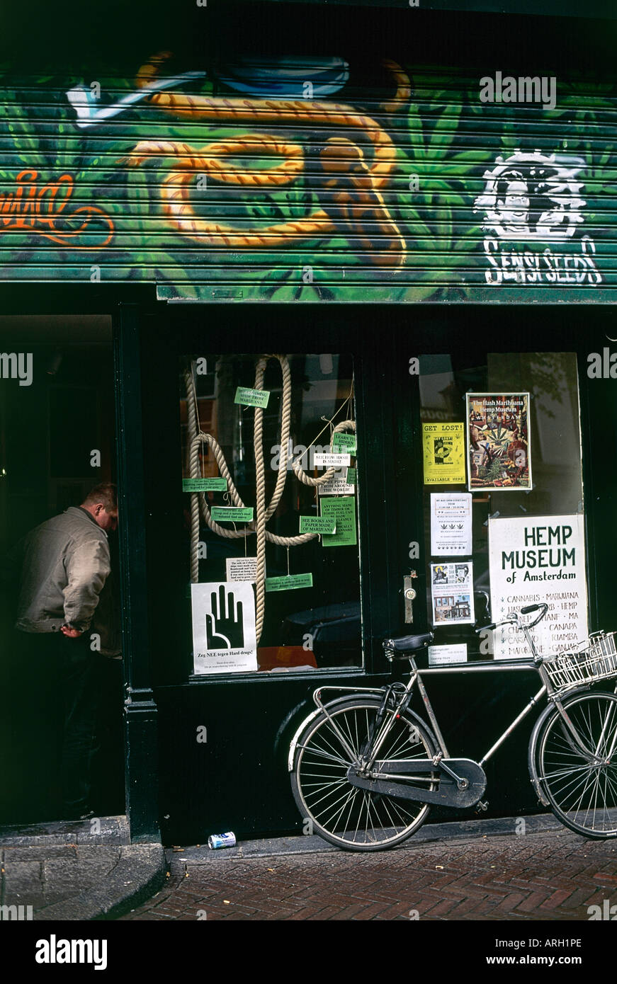 A man studies one of the posters displayed in the entrance of Amsterdam s Hash and Marijuana Museum with a bicycle leaning against the frontage and a painting of a hemp rope amongst marijuana leaves decorating the shutters above Stock Photo
