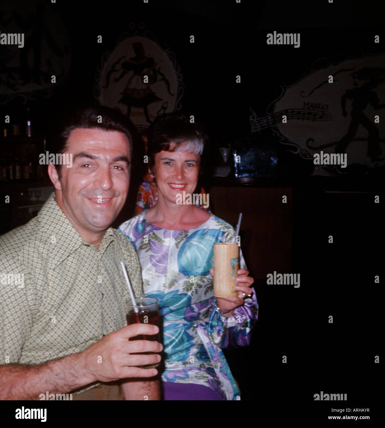 Middle-aged 1970s holiday couple hold drinks and smile at camera. Stock Photo