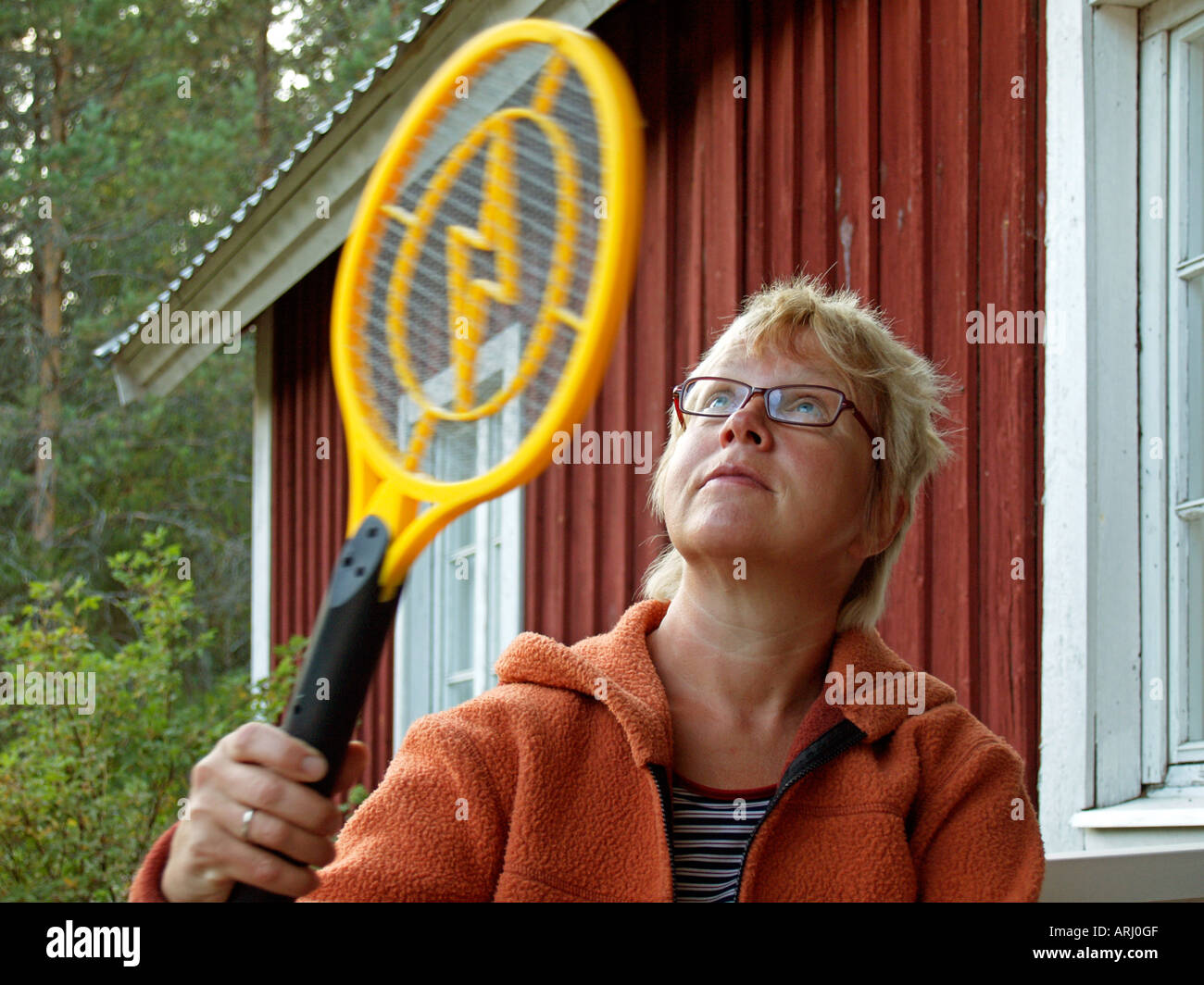woman sitting in front of a red scandinavian timber wooden house trying to catch gnats mosquitos with an electric racket Stock Photo