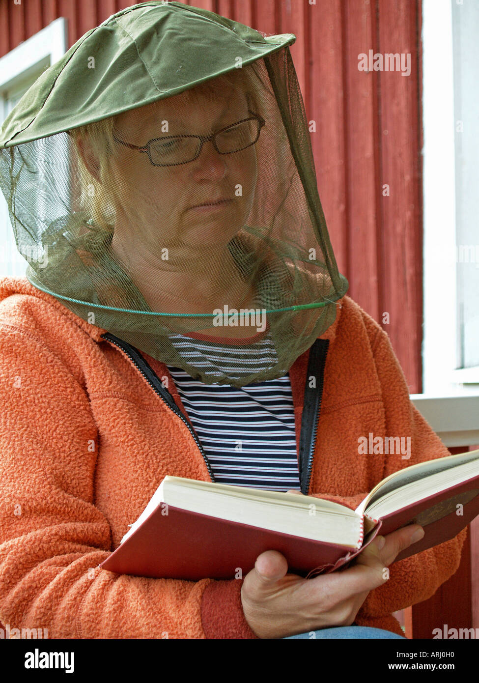 woman with gaze as protection against gnats mosquitos sitting in front of a red scandinavian timber wooden house reading Stock Photo