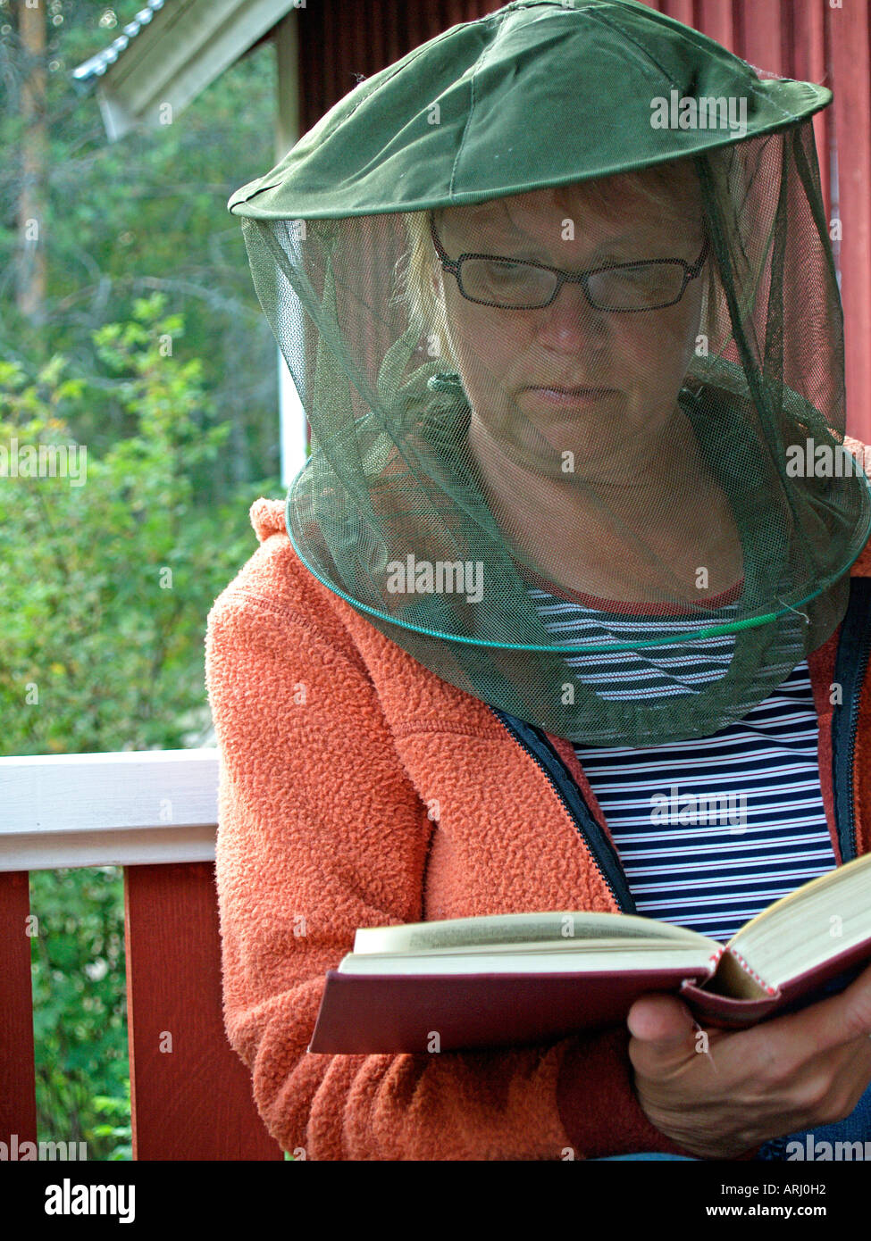 woman with gaze as protection against gnats mosquitos sitting in front of a red scandinavian timber wooden house reading Stock Photo