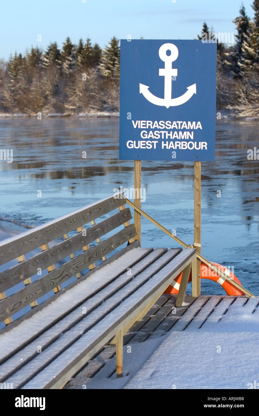 Snowy wooden bench and a multilingual guest harbour sign ( in Finnish , Swedish and  English ) at small boat harbor at Winter , Finland Stock Photo