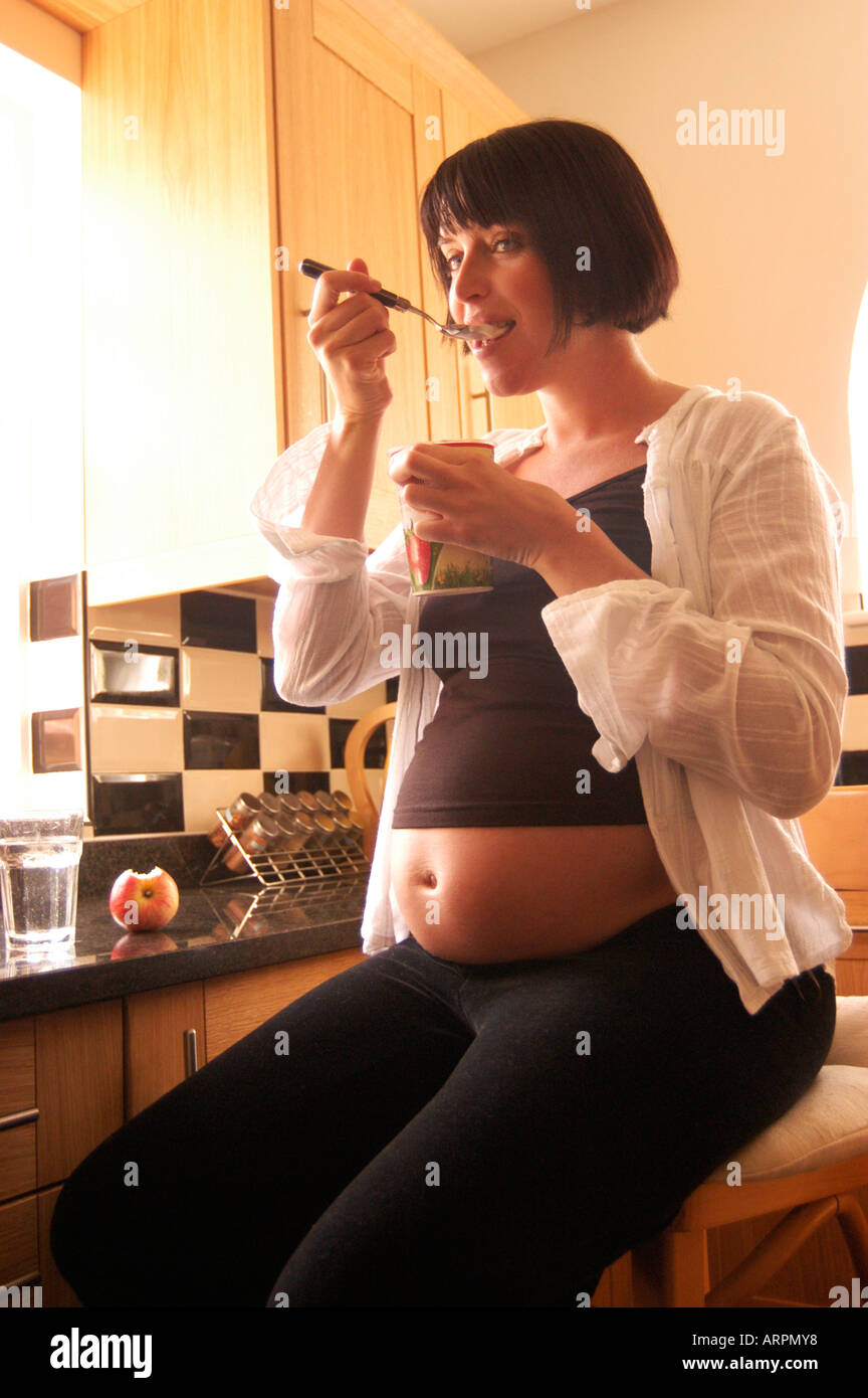 Pregnant woman eating yogurt sitting on a stool in her kitchen Stock