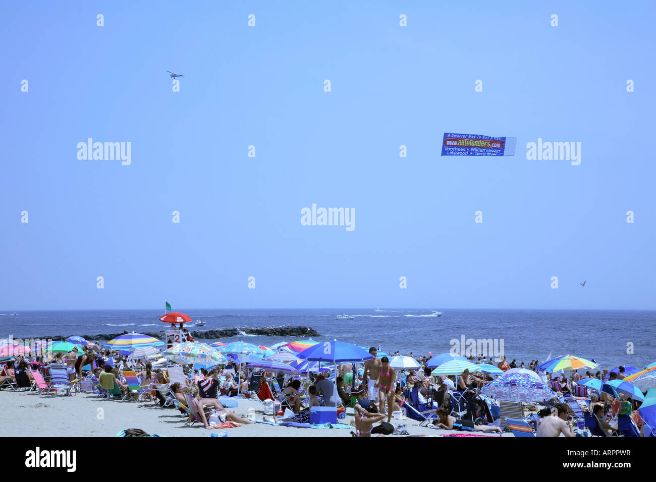 Mass of people amidst large colorful beach umbrellas with small plane dragging advertising banner parallel to shore over ocean. Stock Photo