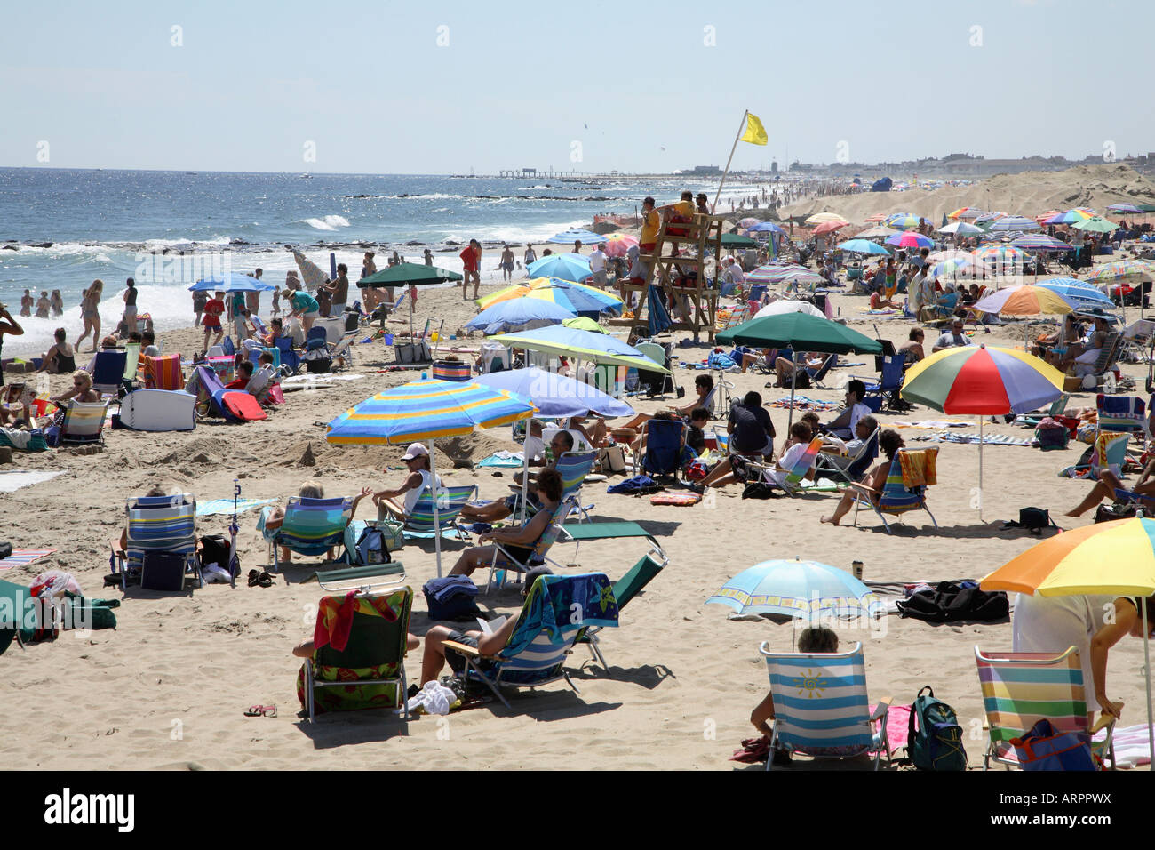Sandy beach with groups of people with chairs, towels, colorful beach umbrellas clustered around lifeguard tower Stock Photo
