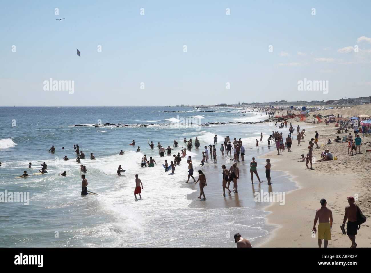 Mass of people playing in shallow surf at oceans edge with plane dragging large advertising banner overhead. Stock Photo