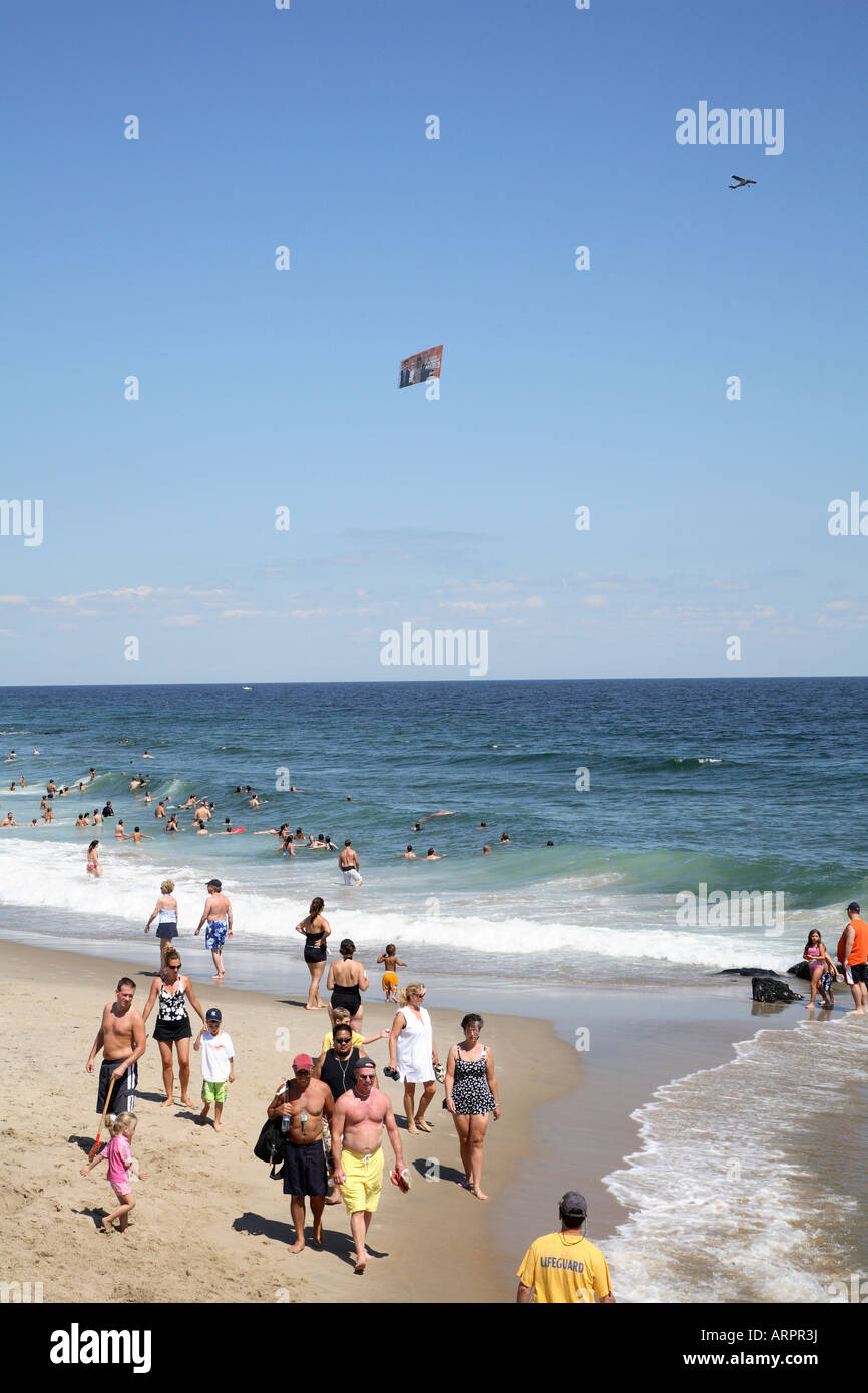 People walking along sandy surf line with wave breaking just behind. Small plane dragging advertising banner above over ocean. Stock Photo