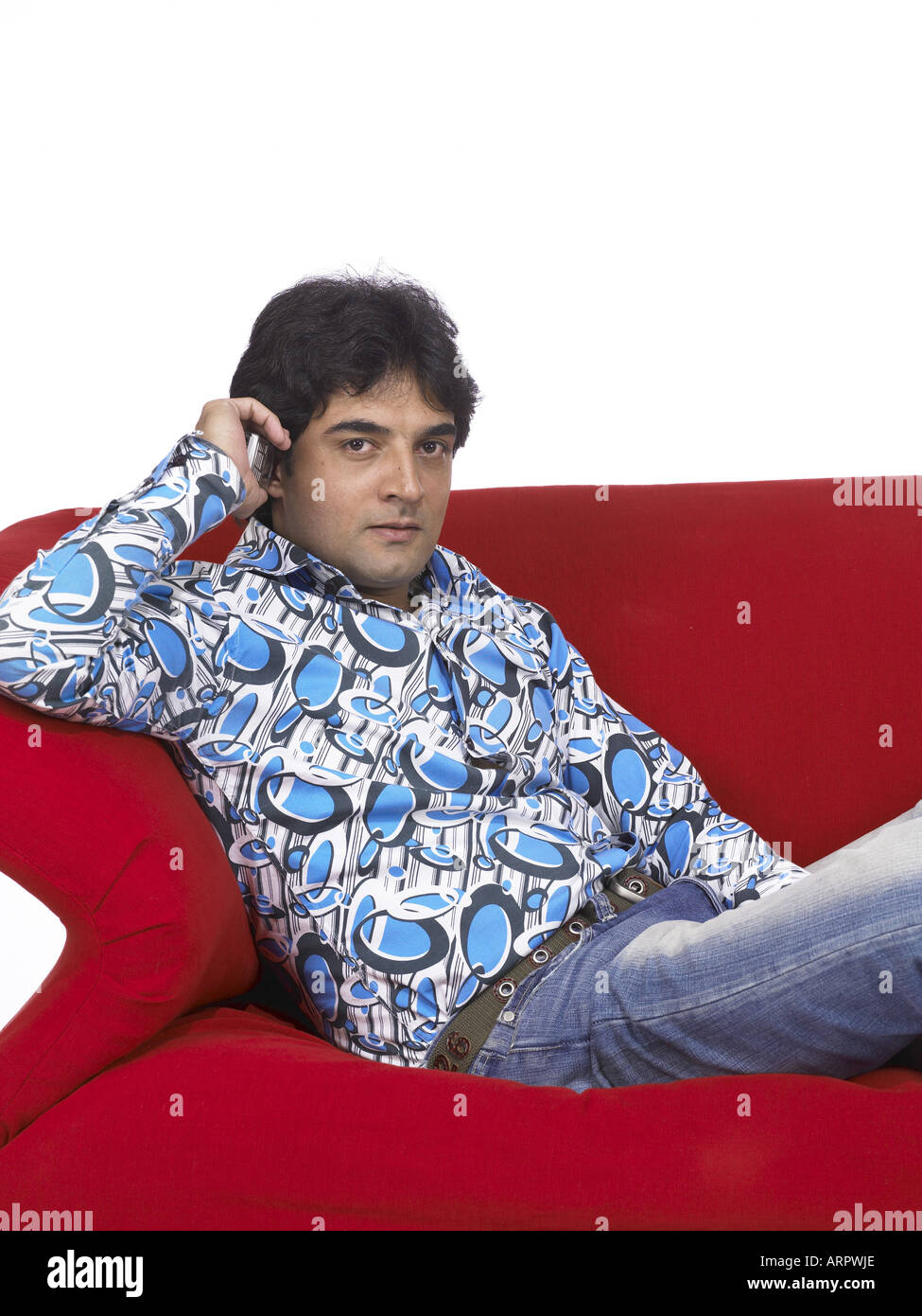VDA200169 South Asian Indian man sitting on sofa talking on mobile looking at camera wearing jeans and shirt MR 701 Stock Photo