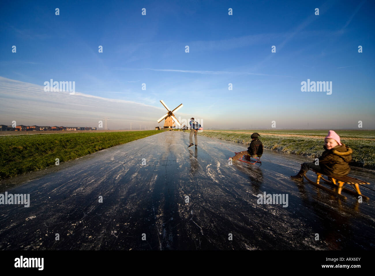 Winter in Holland. Father on skates pulls his two children on toboggans or sleds on the ice towards a windmill. Stock Photo