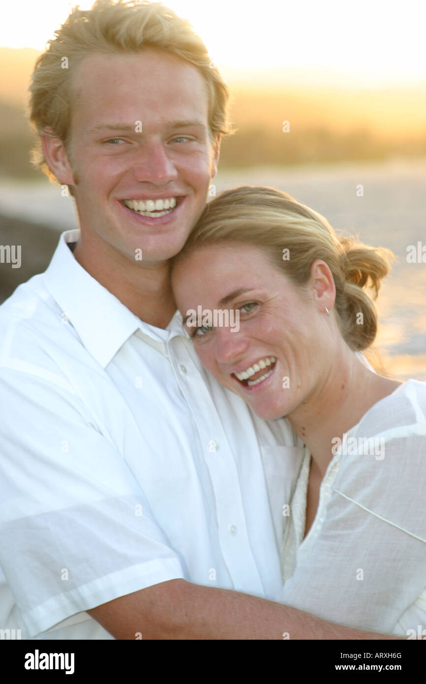 Couple wearing white smile and hug. Golden evening light on the ocean and mountains in the background. Stock Photo