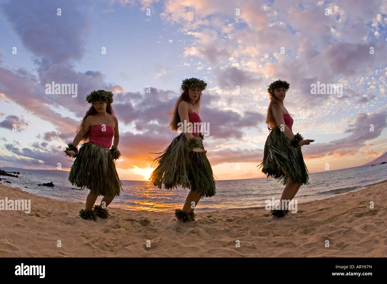 Three hula dancers in ti leaf skirts dance on the beach at sunset at