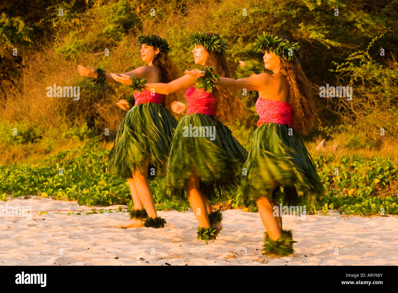 Three hula dancers in ti leaf skirts dance on the beach at sunset at